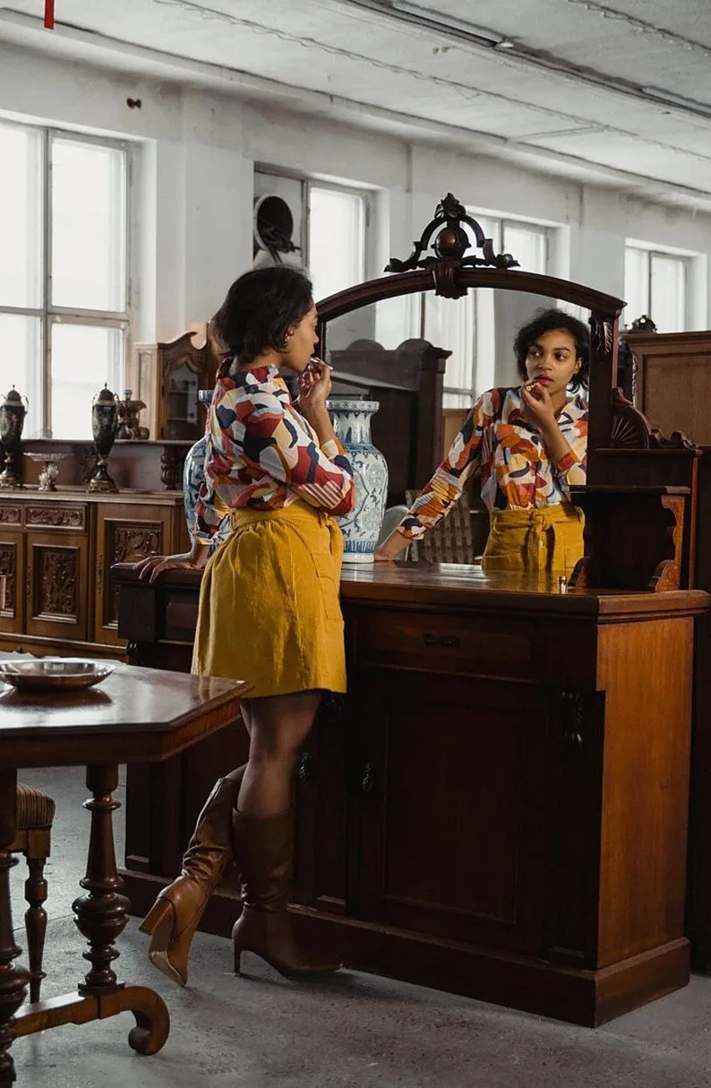 Woman in colorful shirt and boots admiring herself in an antique mirror at a vintage furniture store.