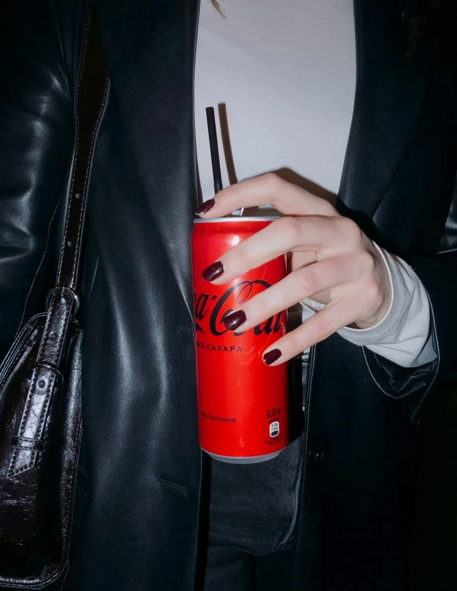 Close-up of a hand holding a red soda can with black straws, against a dark outfit background.